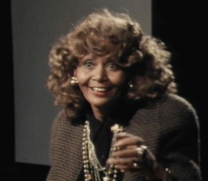 A woman with voluminous, curly hair, dressed in vintage fashion, smiling and accessorized with jewelry.